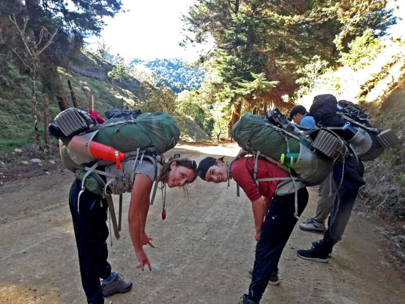 The image shows two people with large backpacks facing each other on a dirt road. They are bending forward, almost touching heads. Behind them, other people with backpacks are walking on the same road. The setting appears to be a mountainous or hilly area with trees and vegetation on either side of the path.